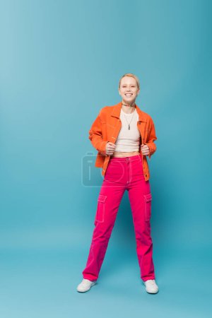 full length of cheerful blonde woman in stylish and colorful outfit standing on blue background