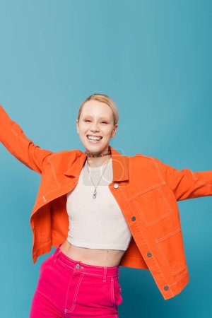 cheerful blonde woman in colorful clothes posing with outstretched hands while smiling at camera isolated on blue