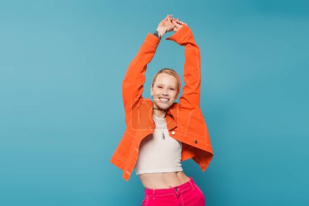 excited woman in colorful clothes posing with raised hands isolated on blue