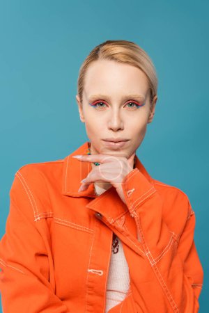 Téléchargez les photos : Portrait of blonde woman with colorful makeup wearing orange jacket and looking at camera isolated on blue - en image libre de droit