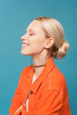 Photo for Side view of joyful blonde woman in colorful beads and orange jacket isolated on blue - Royalty Free Image