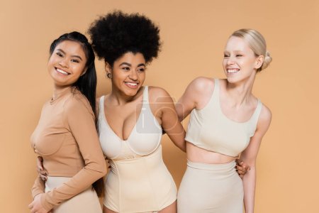 alegre afroamericana mujer en lencería abrazando jóvenes modelos multiétnicos aislados en beige