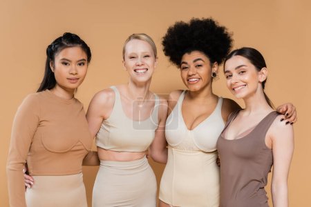 alegres mujeres multiculturales en lencería abrazando y mirando a la cámara aislada en beige