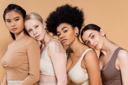 Foto de Young multiethnic women in underwear leaning on each other and looking at camera isolated on beige - Imagen libre de derechos
