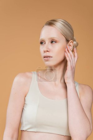 Foto de Young blonde woman in underwear touching face and looking away isolated on beige - Imagen libre de derechos