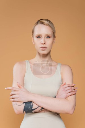 Foto de Vista frontal de mujer rubia tatuada en la parte superior posando con los brazos cruzados y mirando a la cámara aislada en beige - Imagen libre de derechos