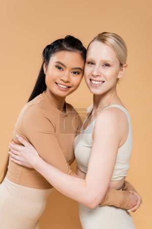 Téléchargez les photos : Blonde and brunette interracial women in underwear embracing and smiling at camera isolated on beige - en image libre de droit