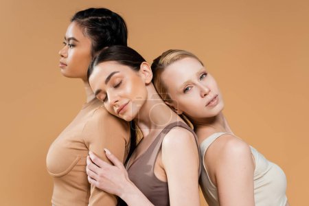Foto de Joven modelo morena posando con los ojos cerrados cerca de las mujeres interracial en ropa interior aislada en beige - Imagen libre de derechos