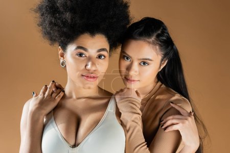 portrait of sensual interracial women in lingerie embracing and looking at camera isolated on beige