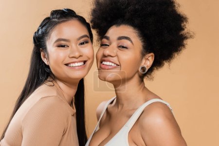 Porträt hübscher afrikanisch-amerikanischer und asiatischer Frauen, die isoliert auf beige Kamera schauen
