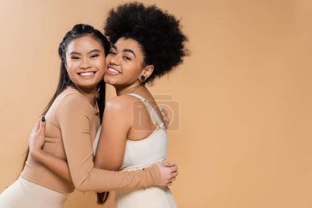 joyful asian and african american women in underwear embracing and looking at camera isolated on beige
