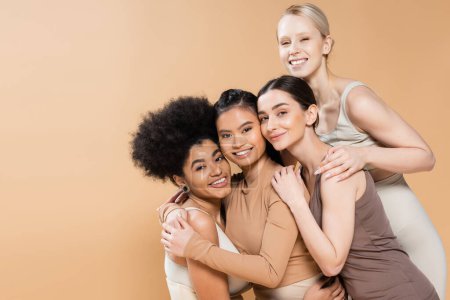 modelos multiétnicos complacidos abrazando y sonriendo a la cámara mientras posan en lencería aislada en beige