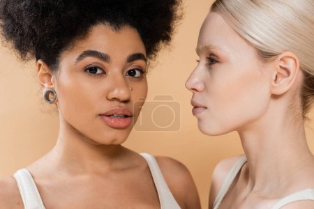 Foto de Retrato de mujer afroamericana bonita mirando a la cámara cerca de modelo rubia joven aislado en beige - Imagen libre de derechos