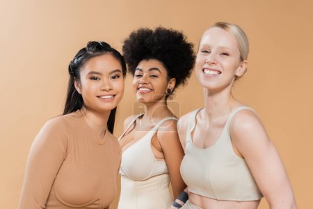 jóvenes y felices mujeres multiétnicas en ropa interior mirando a la cámara aislada en beige