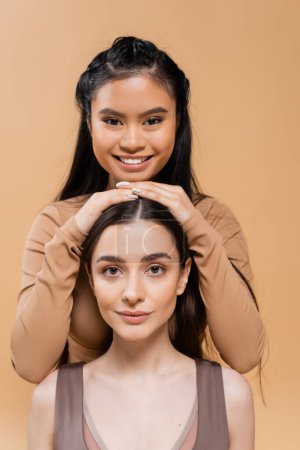 Téléchargez les photos : Cheerful asian woman touching head of young brunette model and looking at camera isolated on beige - en image libre de droit