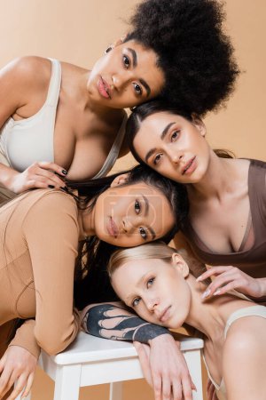 Téléchargez les photos : Young multicultural women in lingerie posing near stool and looking at camera isolated on beige - en image libre de droit