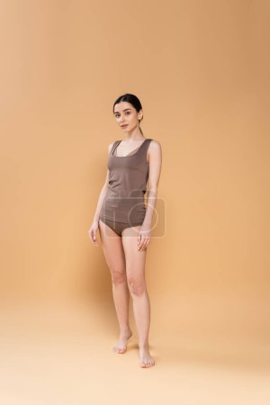 full length of slim barefoot woman in underwear looking at camera while standing on beige background