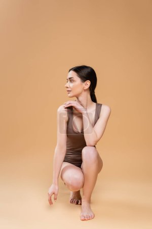 full length of young barefoot woman in underwear looking away while sitting on haunches on beige background
