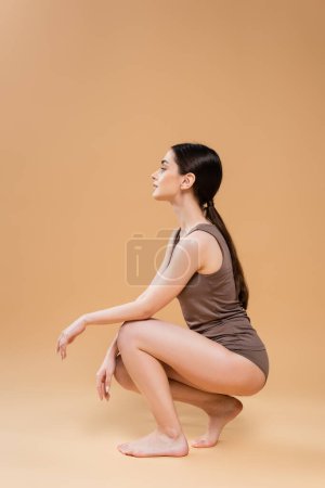 Photo for Side view of slender brunette woman in lingerie posing on haunches on beige background - Royalty Free Image