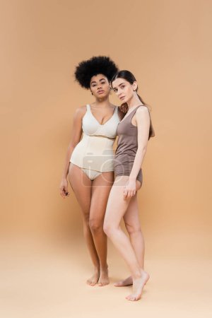 full length of young multiethnic women posing in underwear on beige background