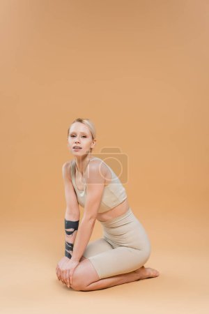 Téléchargez les photos : Pleine longueur de femme blonde tatouée en sous-vêtements assis sur des hanches et regardant la caméra sur fond beige - en image libre de droit