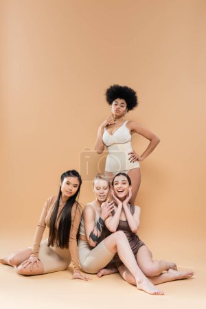 Foto de Full length of sensual african american woman in lingerie posing with hand on hip near multiethnic models sitting on beige background - Imagen libre de derechos