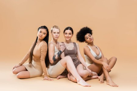 Foto de Full length of young multiethnic women in underwear sitting and looking at camera on beige background - Imagen libre de derechos