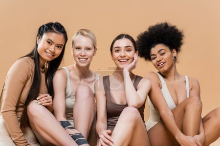 modelos multiculturales satisfechos en ropa interior sentado y sonriendo a la cámara aislado en beige
