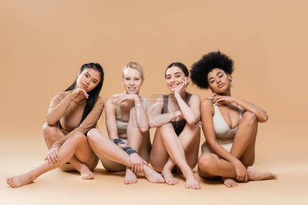 Photo for Full length of pretty multicultural models in underwear sitting with crossed legs on beige background - Royalty Free Image