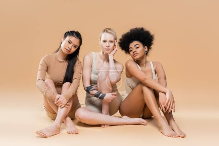 in voller Länge barfuß multikulturelle Frauen in Unterwäsche sitzen und in die Kamera auf beigem Hintergrund schauen