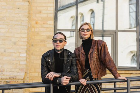 stylish couple in trendy sunglasses and leather jackets standing outdoors 