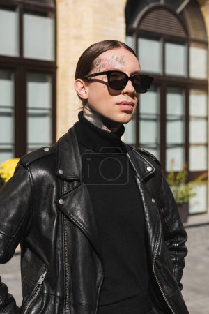 tattooed young man in stylish sunglasses and black leather jacket standing on urban street 