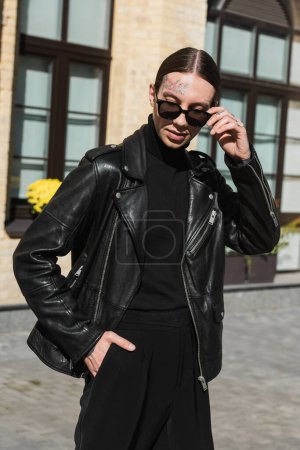 tattooed young man adjusting stylish sunglasses and standing with hand in pocket on urban street 
