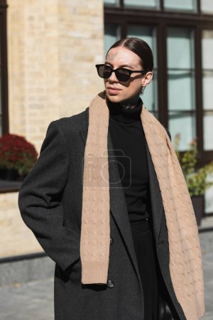 Foto de Tattooed young man in coat and stylish sunglasses standing with hand in pocket and looking away - Imagen libre de derechos