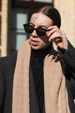 tattooed young man in coat and scarf adjusting stylish sunglasses