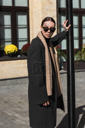 Tätowierter junger Mann in Mantel und stylischer Sonnenbrille steht in der Nähe von Straßenmasten auf der städtischen Straße 