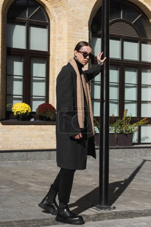 in voller Länge tätowierter junger Mann in Mantel und stylischer Sonnenbrille, der in der Nähe von Straßenmasten auf der städtischen Straße steht 
