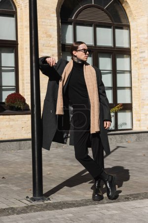 Ganzkörpertätowierter Mann in trendigem Mantel und stylischer Sonnenbrille, der mit überkreuzten Beinen auf der Straße steht 