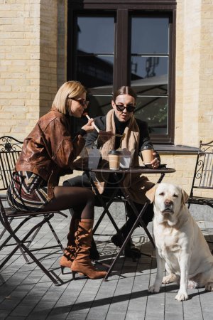 stylisches junges Paar isst asiatisches Essen in der Nähe von Kaffee, um mit Labrador auf dem Bistrotisch zu sitzen 