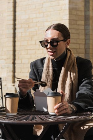 stylish tattooed man in sunglasses and coat holding chopsticks while eating asian food 