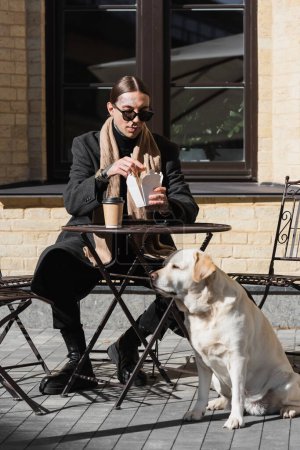Ganzkörpertätowierter Mann mit Sonnenbrille hält Essstäbchen in der Hand, während er asiatisches Essen in der Nähe eines Hundes in einem Outdoor-Café isst 