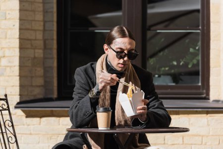Téléchargez les photos : Homme tatoué dans des lunettes de soleil et manteau tenant des baguettes tout en mangeant des aliments asiatiques près de tasse en papier sur la table de bistrot - en image libre de droit
