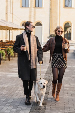 full length of happy couple in stylish sunglasses holding paper cups while walking with dog 