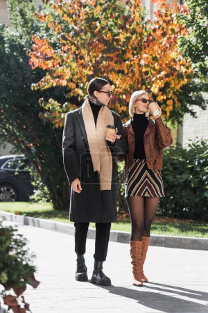 full length of stylish couple in sunglasses holding paper cups while walking outside 
