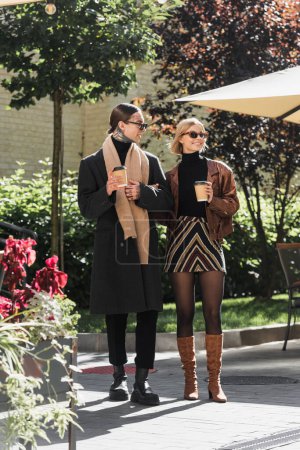toute la longueur du couple élégant dans des lunettes de soleil tenant des tasses en papier tout en se tenant près du café extérieur 