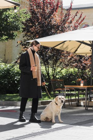 Ganzkörpertätowierter Mann mit trendiger Sonnenbrille, der Pappbecher hält, während er mit Labrador in der Nähe eines Cafés spaziert 