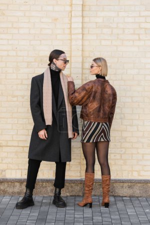 full length of stylish woman in sunglasses standing with tattooed man near brick wall  