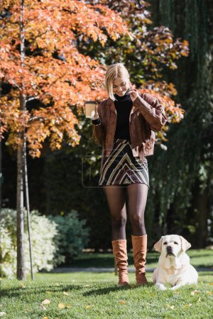 full length of happy blonde woman holding paper cup and walking with labrador in park 