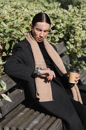 young tattooed man in coat and scarf holding takeaway drink while looking at wristwatch and sitting on bench 