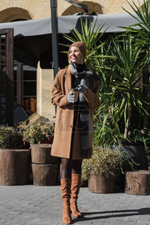 Foto de Longitud completa de mujer rubia feliz en abrigo beige y sombrero sosteniendo taza de papel con café para llevar - Imagen libre de derechos